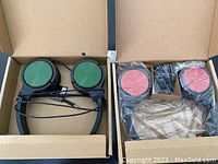 Two pairs of AmazonBasics on-ear headphones in open cardboard boxes, one with green ear pads and another with red ear pads.