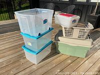 Overview of seven assorted plastic storage bins and laundry basket of different sizes and styles on wooden deck.
