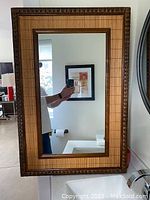 Full view of rectangular rattan framed mirror hung on wall reflecting part of room and window.