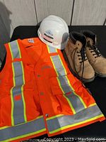 Photo of large orange safety vest with reflective stripes, white hard hat, and tan steel toed boots on black surface