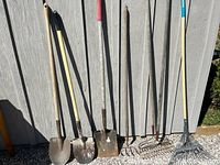 Full view of seven garden tools including three shovels, two garden forks, and two rakes leaning against a wooden shed.