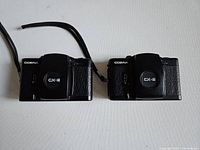 Front view of two Cosina CX-2 compact 35mm cameras showing lens caps with brand and model markings and one camera with a wrist strap attached.