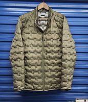 Front view of a men's Columbia jacket in green hanging on a hanger inside a storage unit. Shows full front zipper and quilted raised geometric pattern.