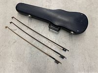 Black hard violin case closed with 3 vintage violin bows laid in front of it on a light-colored surface.