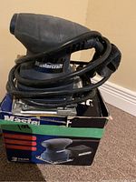 Front view of Mastercraft 1/4 sheet sander with power cord coiled around it, sitting on original box.