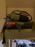 Ryobi angle grinder resting on its original box, showing the full tool from side profile with power cord attached.