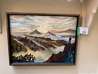 Framed acrylic painting measuring 25 by 19 inches depicting a scenic mountain landscape with mist and clouds, showing an individual standing on a cliff edge.