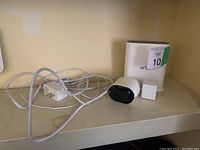 Photo showing white charging station, power adapter with cable, small white accessory, and black Arlo camera on shelf
