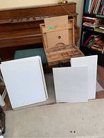 Wooden art box open with compartments, several blank white canvases and canvases with faint pencil sketches arranged in front