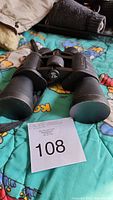 Black Outbound binoculars lying flat showing front large objective lenses and focus wheel, placed on colorful blanket with auction lot number 108.