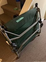 Side view of folded green fabric camping bed with metal frame, shown indoors on carpeted floor next to stairs.