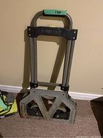 Foldable dolly leaning against a wall, showing metal frame, foam handle grip with '150' marking, wheels and rusted base.