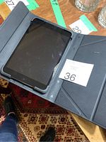 iPad inside a black folding protective case, displaying the screen and home button, resting on a patterned carpet.