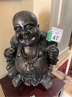 Photo shows a Laughing Buddha statue made of dark material, placed indoors on a wooden surface.