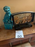 Photo of turquoise ceramic guardian lion statue and wood-framed glass carved scene on a wooden surface.