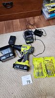 Ryobi cordless drill with attached battery, an additional battery, charger, and yellow case containing drill bits spread on carpet.