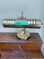 Front view showing cylindrical brass shade with horizontal slots, attached to brass pedestal base on wood surface.