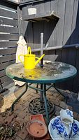 Outdoor round metal table with glass top, yellow watering can placed on top, black metal umbrella stand attached to the center of the table. Surface has dried leaves and dirt.