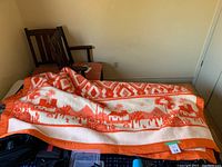 Orange and white vintage blanket partially unfolded on a bed with visible geometric and scenic patterns.