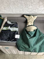Two dog coats displayed on a bench — black with reflective stripe and green with beige trim.