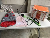 View of two dog jackets, a black leash with green handle, a white spotted ball thrower dog toy, and an orange trim soft-sided portable dog crate on a table.