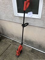 Full view of the Black & Decker electric grass shear showing the orange body, black shaft, and cutting head at the bottom leaning against an exterior wall.