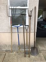 Full view of six garden tools leaning against wall, showing all handles and working ends.