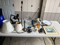 Complete lot of household items displayed on a table including lamps, figurines, glass pitchers, and artwork.