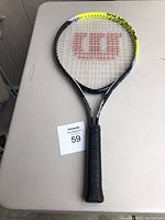 Top view of a black, white, and yellow Wilson kids tennis racket with red Wilson logo on strings, placed on a white surface with a lot tag visible