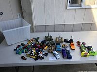 Overview of all children's toys including toy vehicles, remote controls, spinning tops, and large plastic bin on a white table.