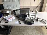 Multiple kitchen appliances and cookware items on a white table in a garage setting.