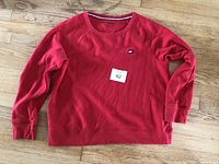 Front view of a red ladies' shirt by Tommy Hilfiger laid flat on a wooden floor. Shirt size indicated as large.