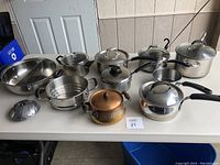 Photo showing ten cooking pots of various materials including stainless steel and copper arranged on a white table.