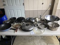 Overview of assorted pans and kitchen supplies on a folding table including frying pans, colander, mixing bowls, and various lids.