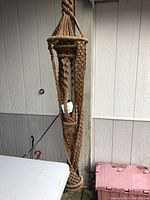 Photo of the woven hanging plant holder suspended against a beige wall near a white table and pink storage container.