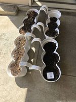 Four white plastic plant boxes arranged in two rows on concrete. Filled with soil and dried leaves, each has three circular connected planting sections and handles on each end.