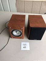 Pair of wooden Curtis bookshelf speakers, one without grill showing silver driver, the other with black grill and Curtis logo.