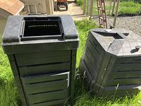 Two black compost containers on grass; one tall with one lid on side, one smaller with hexagonal top