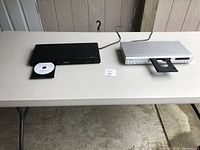 Black Philips DVD player and silver unbranded DVD player both on plastic table with trays open.