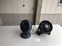 Two black table fans side by side on a white table, one Sunbeam and one Bissell.
