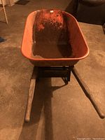Front view of orange metal wheelbarrow showing tub and wooden handles on concrete floor.
