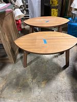 Two elliptical leaf-shaped wooden tables, different heights, standing on floor in a warehouse setting