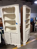 Two white corner cabinets side by side showing open shelving and lower cabinets