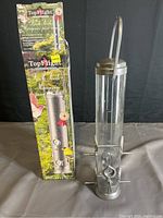Photo of the complete bird feeder next to its original box. The feeder is metallic with a hanging handle and multiple feeding ports around the base.