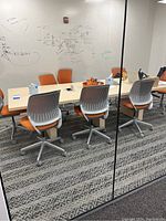 Rectangular conference room table with orange swivel chairs around it, table center has an electrical outlet module. Glass walls reflect the room.