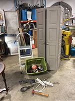 Photo showing entire lot with beige plastic storage cabinet, small metal step ladder, green plastic bin filled with hand tools and supplies, hammer, light, and cords on concrete floor in a workshop setting.