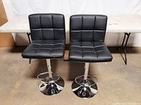 Front view of a pair of black adjustable bar stools with tufted cushions and chrome bases with footrests, arranged side by side.