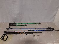 Three metal collapsible hiking poles, two green Kabuta and one blue Cascade, laid flat showing extended sections and wrist straps.