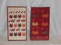 Photo showing two vertical framed needlepoint artworks side by side. The left piece has rows of animals in red, black, and orange shades on a beige background. The right piece depicts stylized birds on a red fabric background.