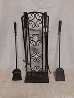 Front view of the wrought iron fireplace tool rack with five tools on and around it, including broom, shovel, poker, and tongs. The rack features swirled decorative accents and a thermometer in center.